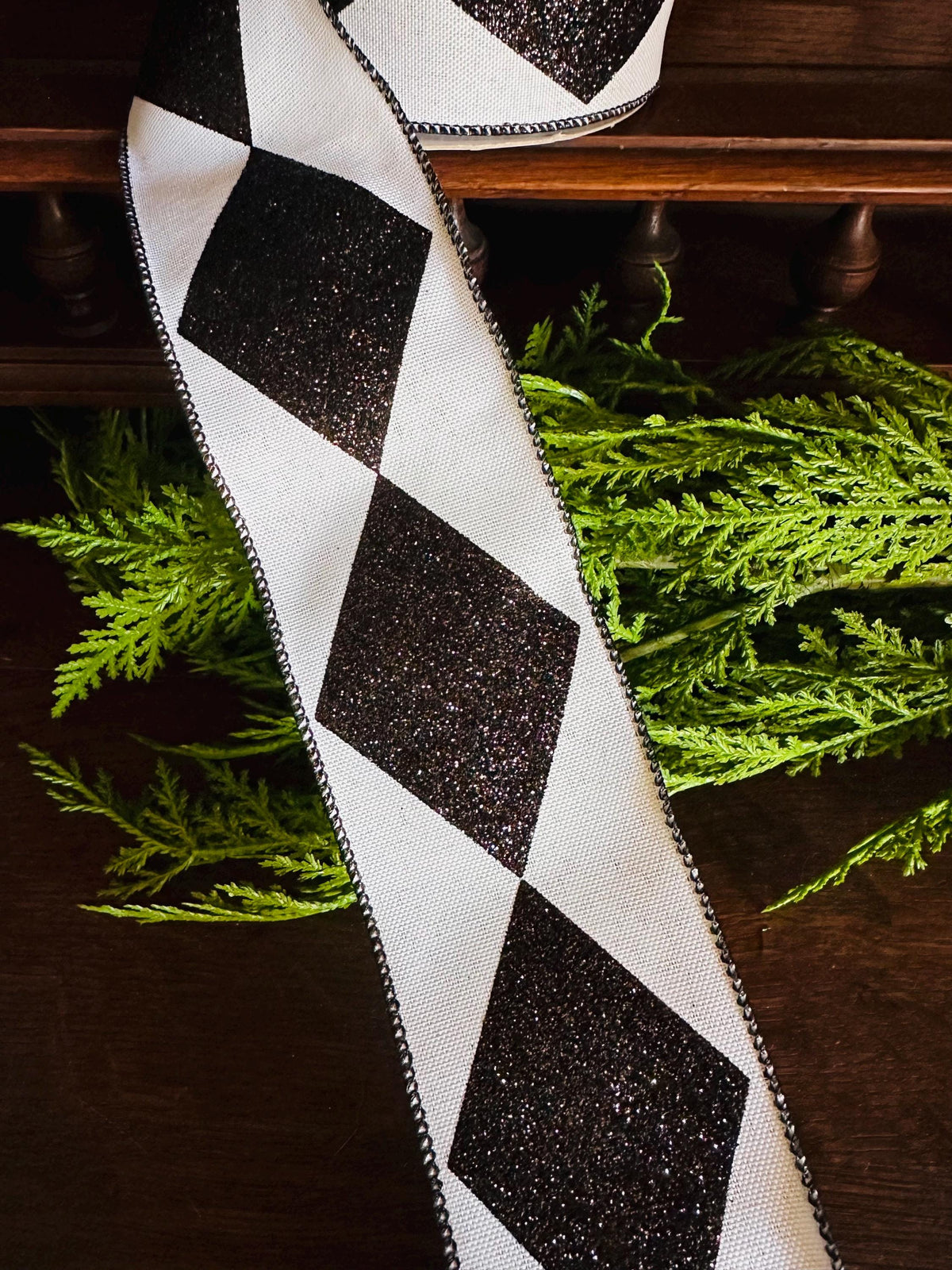 Black and white diamond patterned ribbon with black glitter. The ribbon is shown on a dark brown surface next to green foliage. The ribbon has a wired edge and is suitable for crafting, gift wrapping, and decorating.