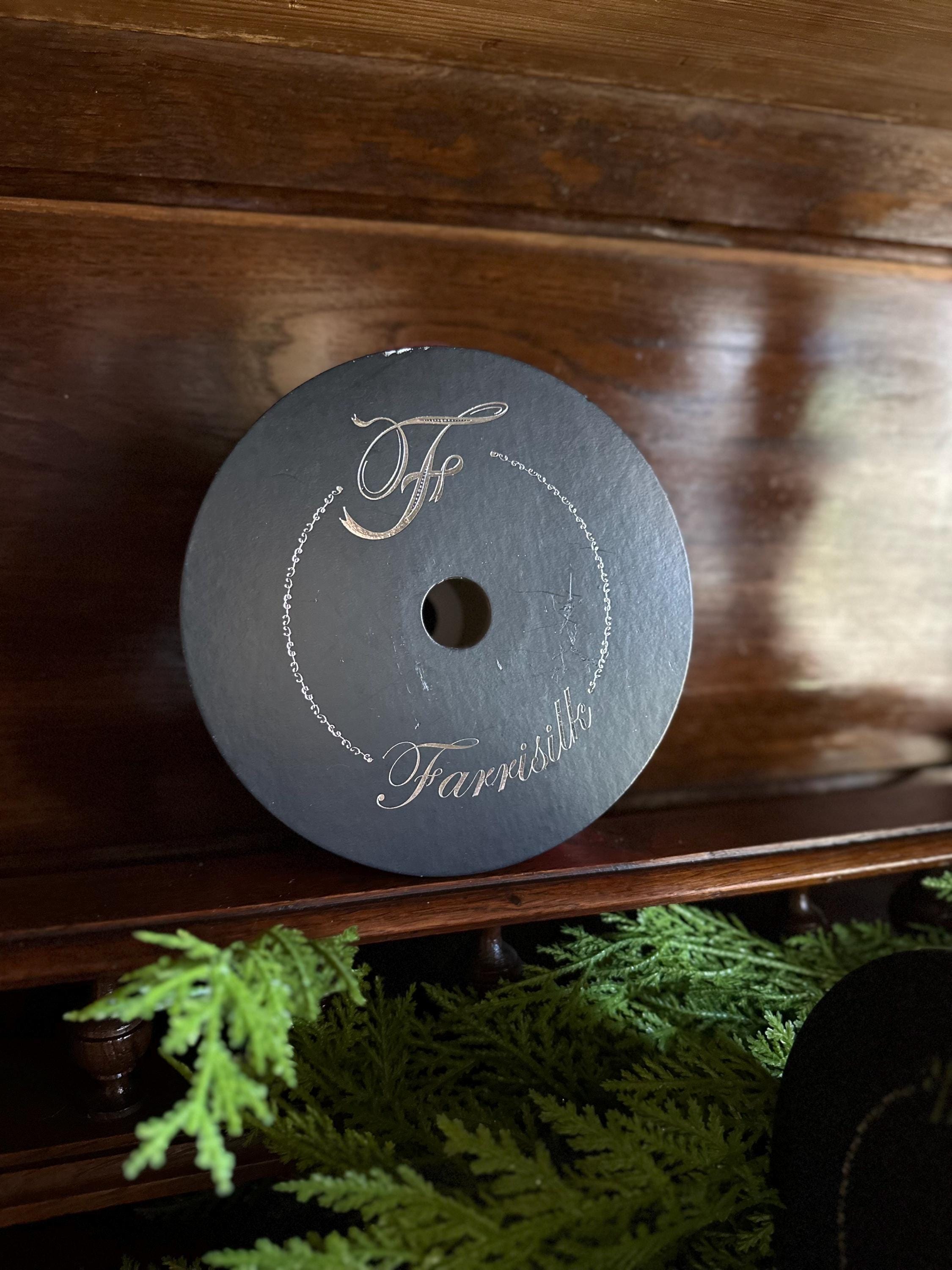 A round black slate keepsake box lid with silver engraved script reading 'Tf' and 'Farrish' on a dark wooden shelf with green foliage at the bottom.