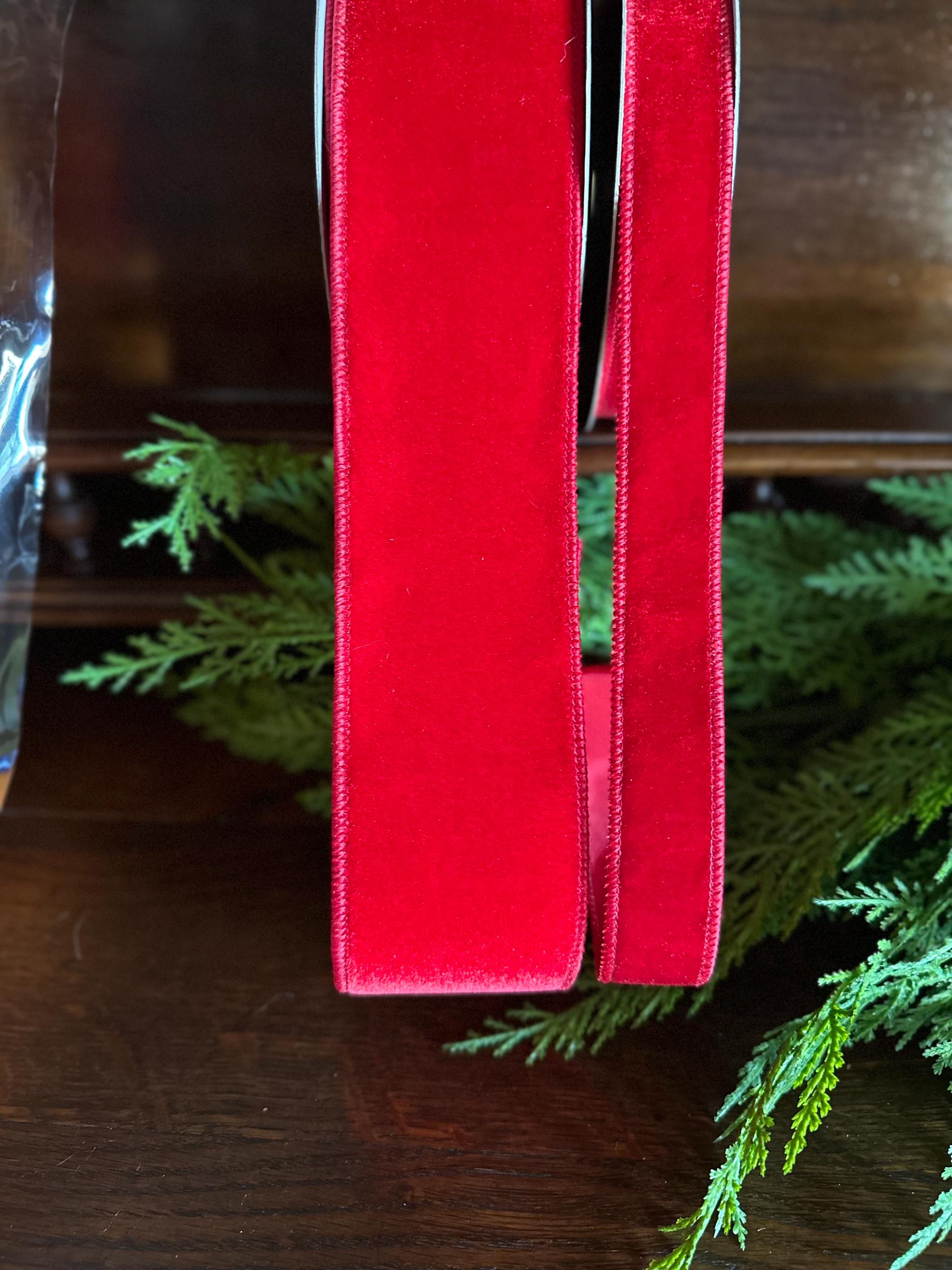 A close-up of a bright red velvet ribbon standing vertically on a dark wooden surface, with green pine branches placed around it.