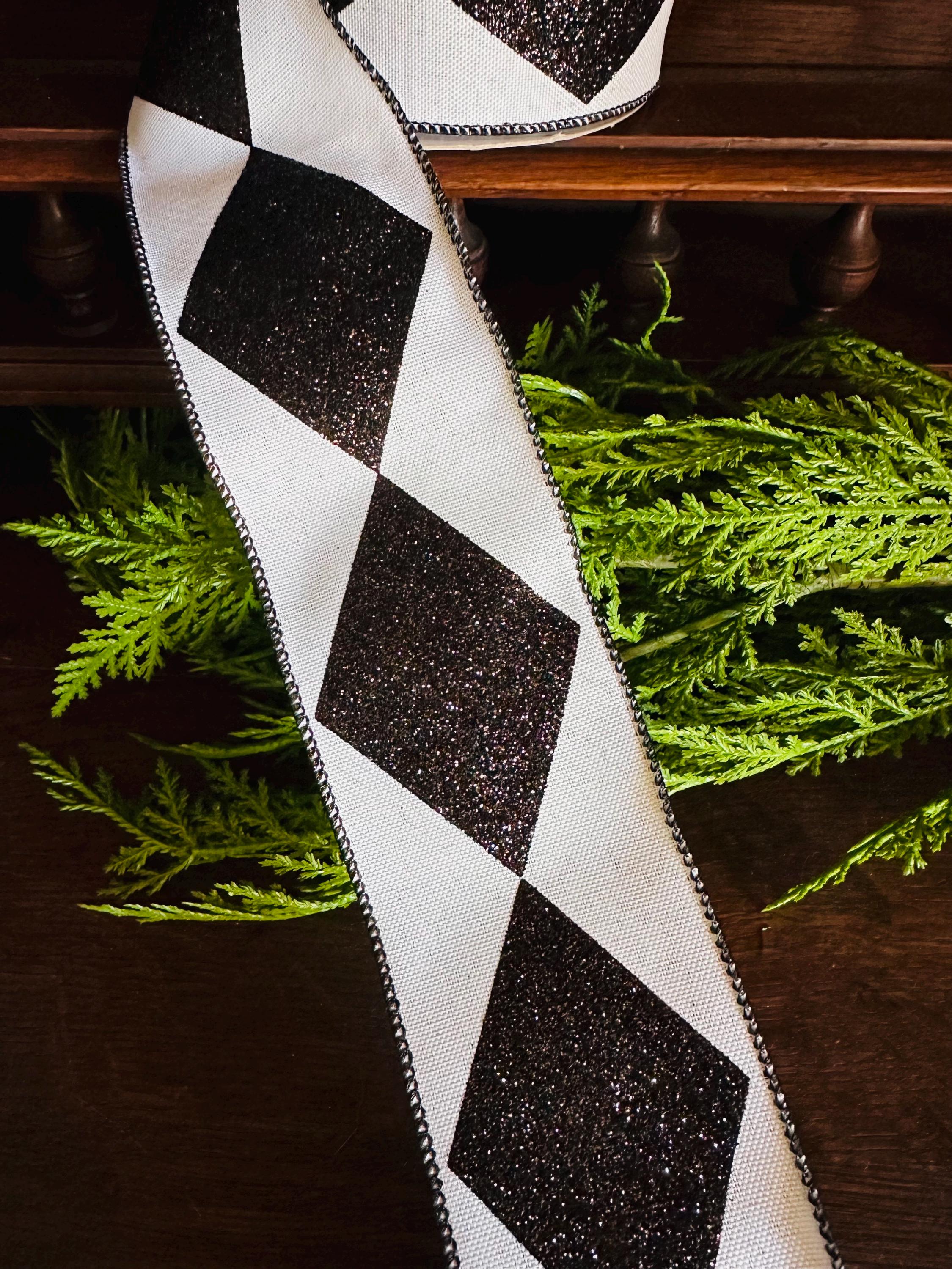 Black and white diamond patterned ribbon with black glitter. The ribbon is shown on a dark brown surface next to green foliage.  The ribbon has a wired edge and is suitable for crafting, gift wrapping, and decorating.