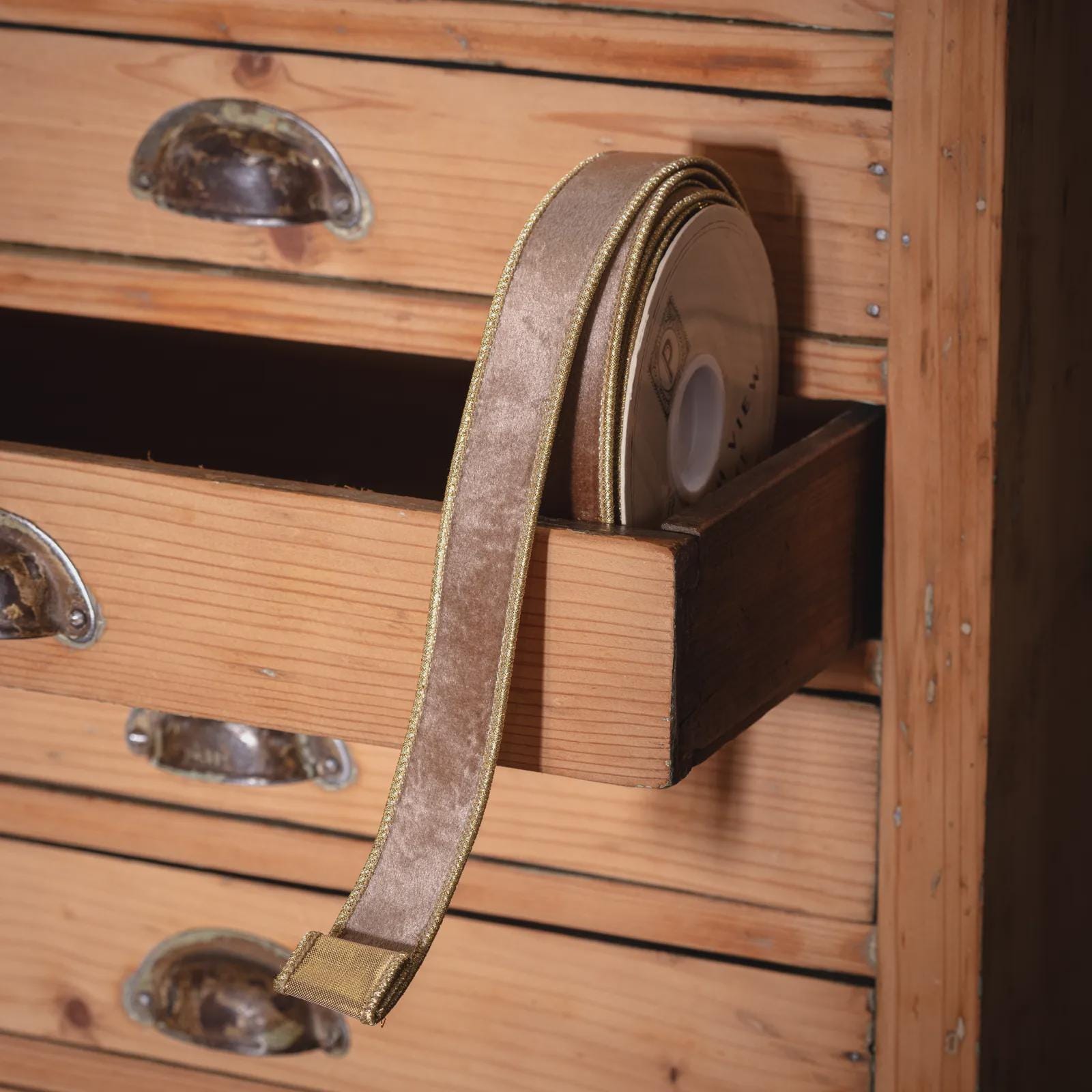 This is a roll of emerald green velvet ribbon displayed against wooden drawers, suggesting a vintage or rustic aesthetic. The ribbon appears to be of high quality with a luxurious sheen, making it ideal for gift wrapping, wedding decorations, Christmas crafts, holiday decor, or special occasion embellishments. This type of ribbon would appeal to crafters, wedding planners, and those looking for premium gift wrapping supplies.