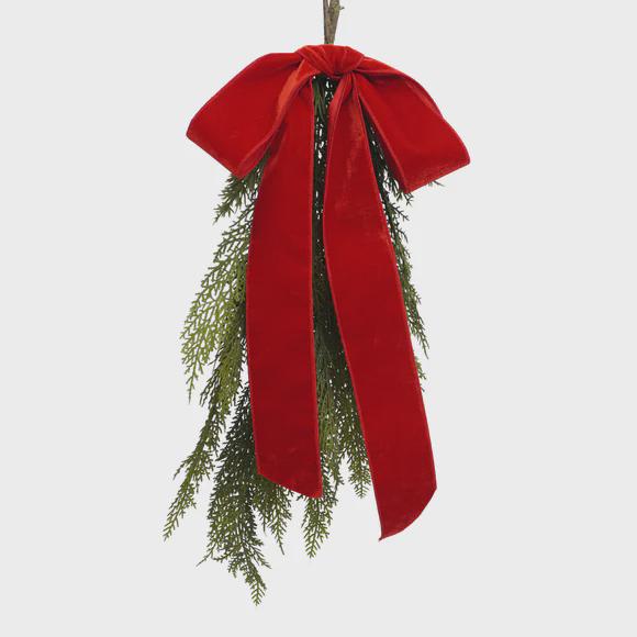 A festive hanging garland featuring green pine and cedar branches tied at the top with a large, wide red velvet bow, shown against a solid light gray background.