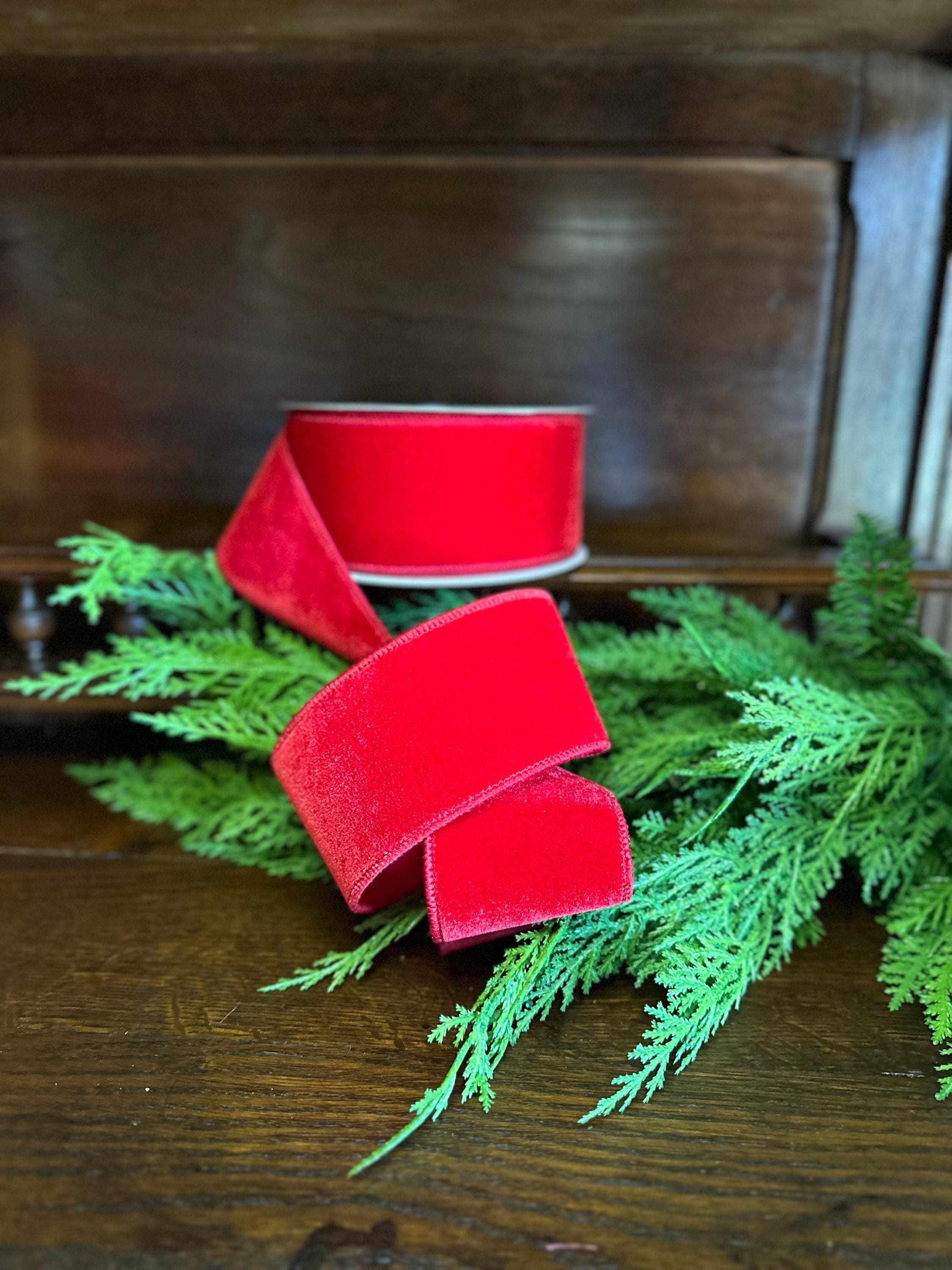 A roll of bright red velvet ribbon draped over green pine branches on a dark wooden surface.