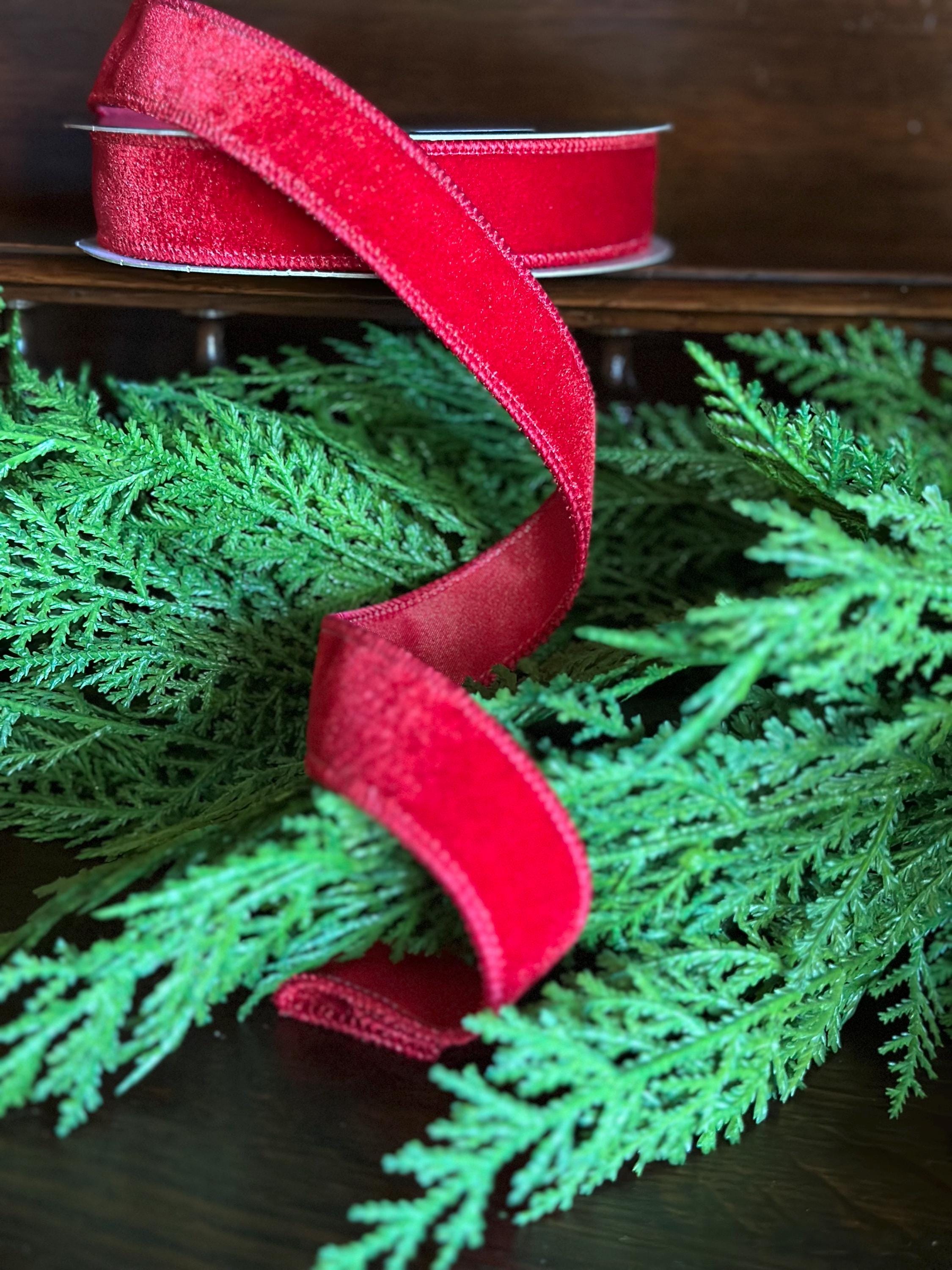 A spool of bright red velvet ribbon draped over green pine branches on a dark wooden surface.