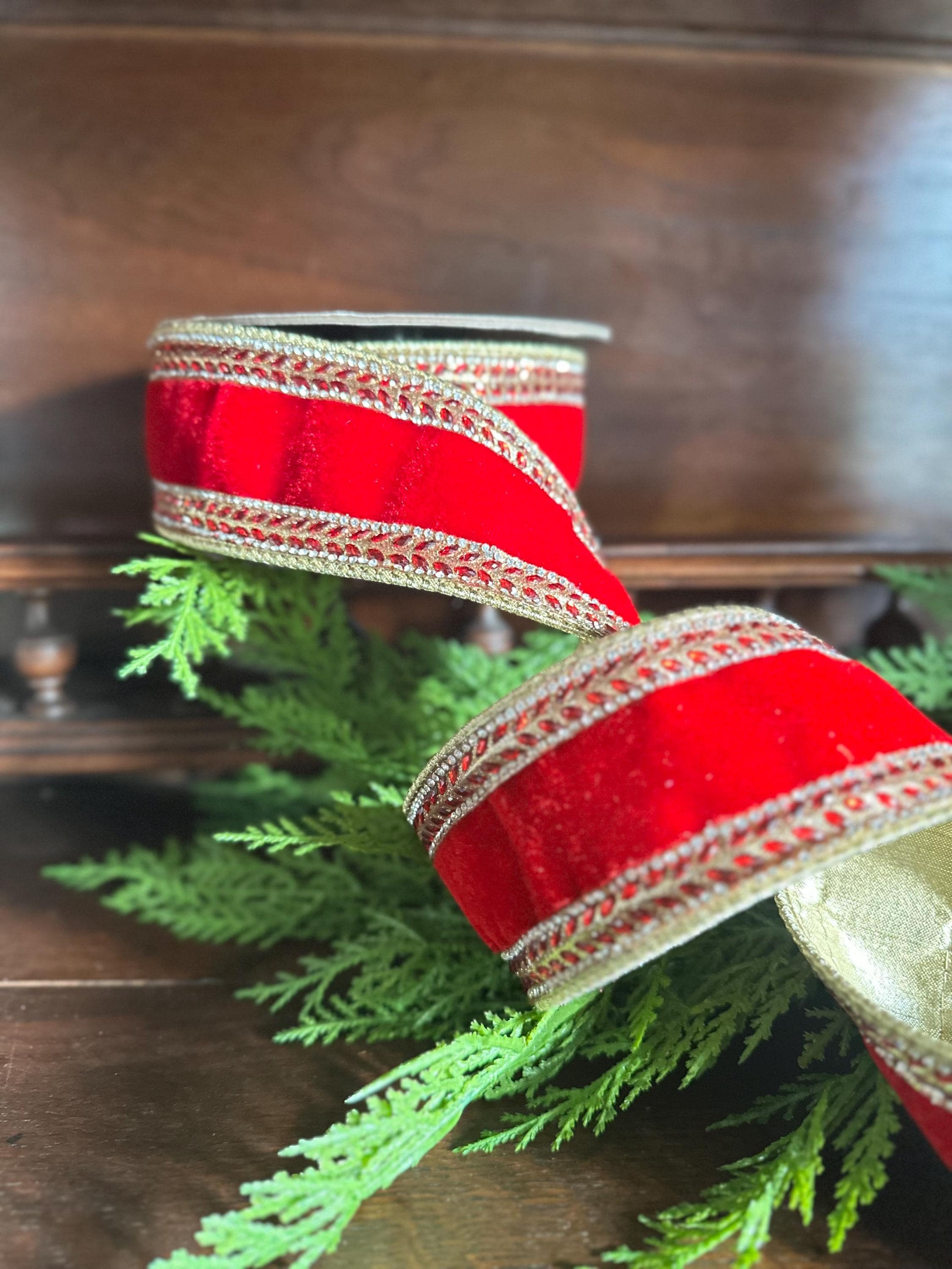 May include: Two rolls of red velvet ribbon with gold trim. The ribbon is decorated with a gold beaded edge. The ribbon is resting on a bed of green pine needles, with a dark wooden surface in the background. The ribbon is likely for holiday or craft use.
