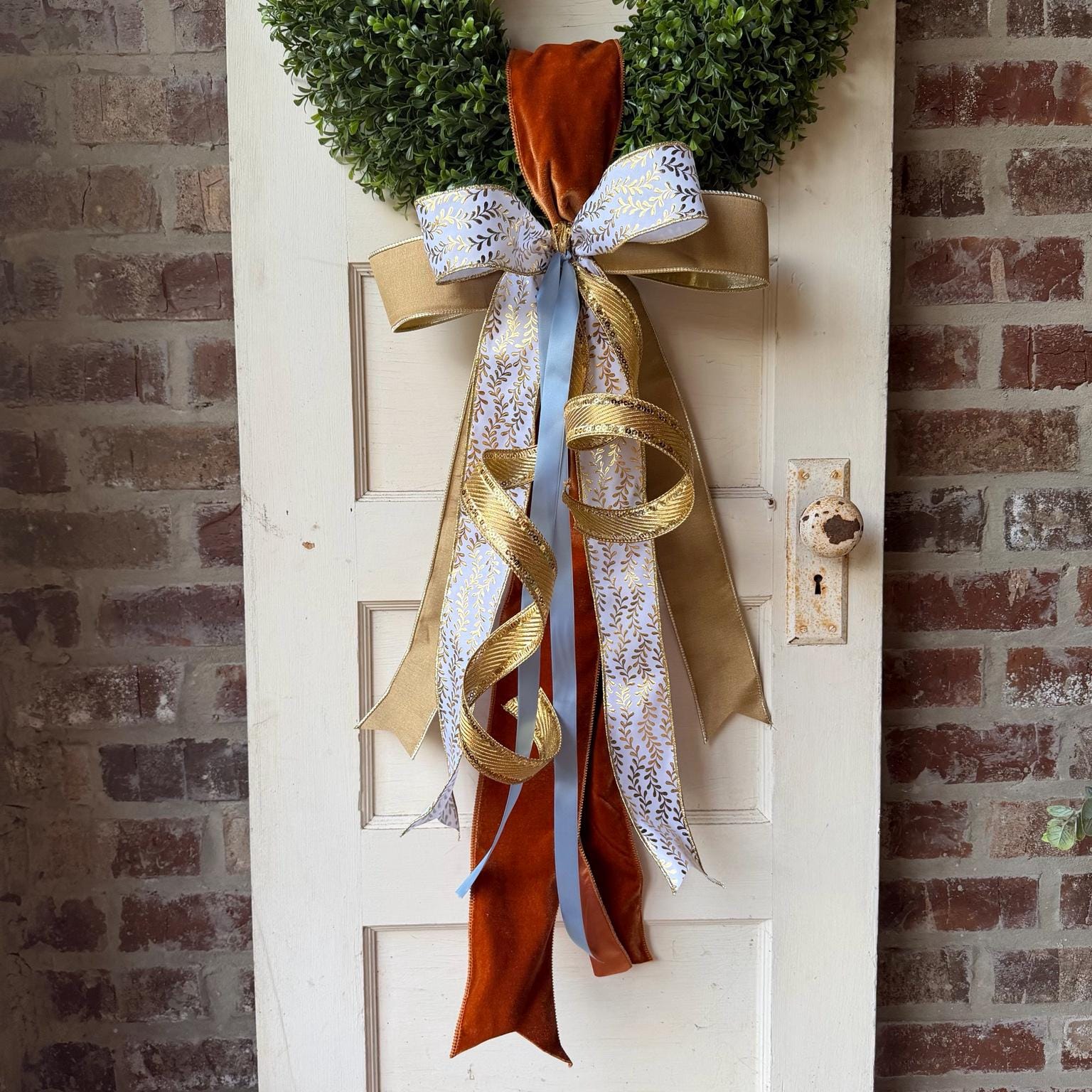 Luxury fall wreath sash with burnt orange velvet, gold ribbon, white metallic leaf ribbon and soft blue satin displayed on a green boxwood wreath